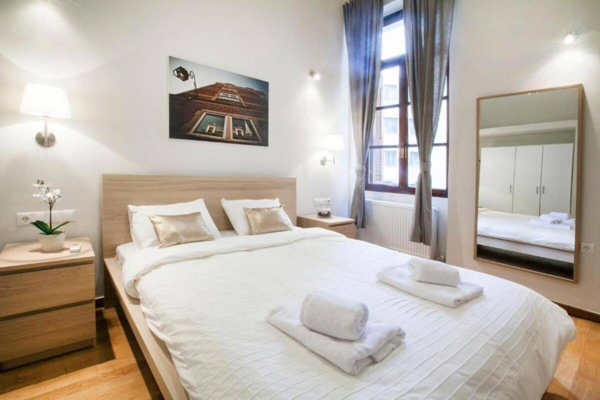 Ein Hotelzimmer mit einem großen Bett, Nachttischen, einer Lampe, einem Blumentopf, einem Fenster mit Vorhängen, einem Wandspiegel und einem Gemälde.