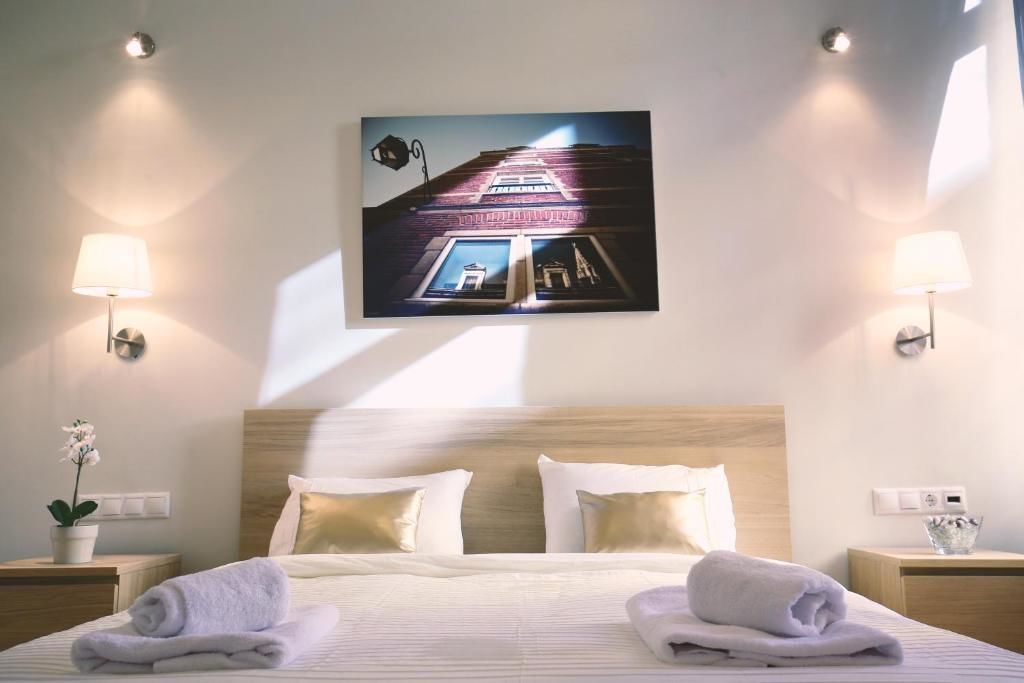 A hotel room with a bed, two nightstands, a wall-mounted lamp, a potted plant, and a painting of a building.