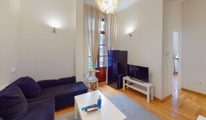 A living room with a dark-colored sofa, a white coffee table with books and a water bottle, a flat-screen TV on a stand, and a wooden floor. - Central Apartments in Psiri (Athens)