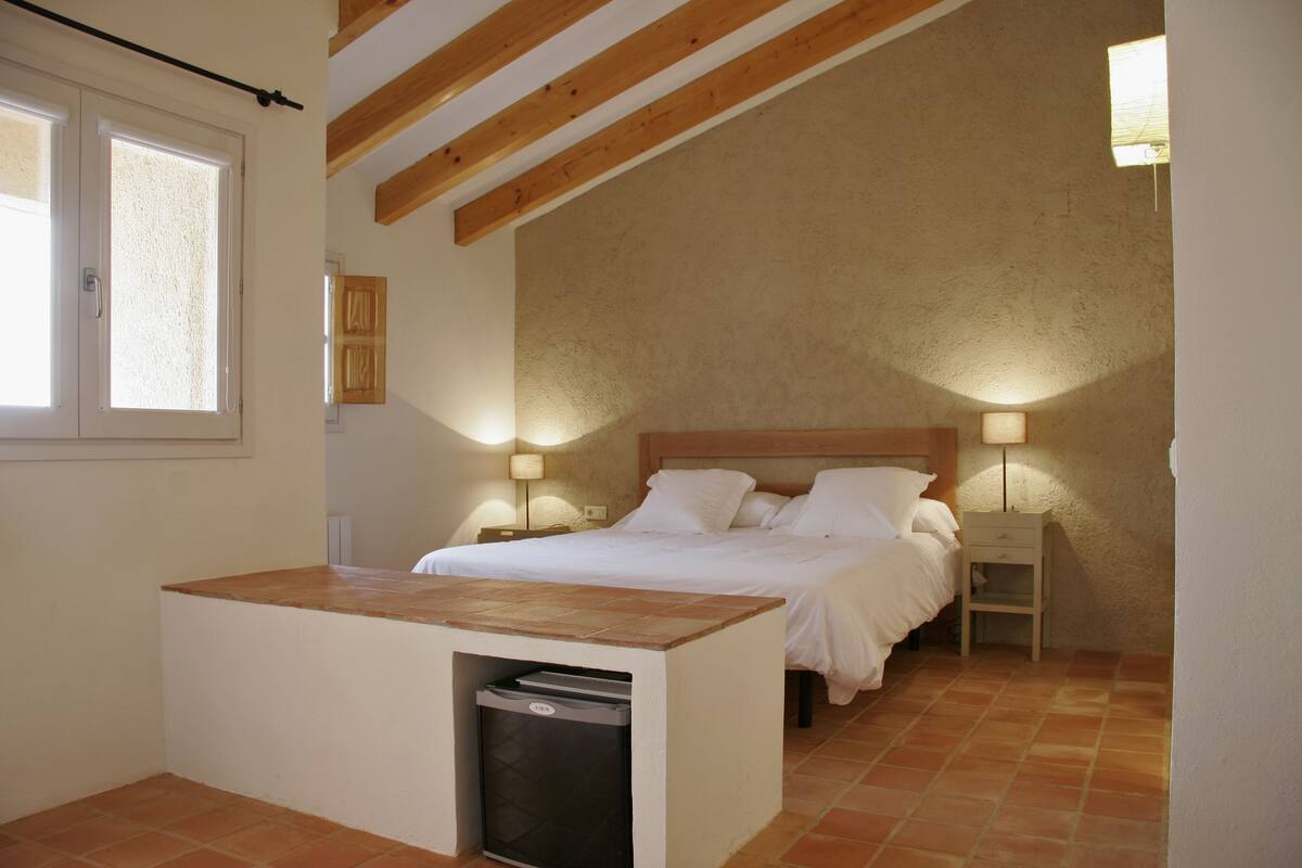 A bedroom with a bed, bedside tables, a small desk with a trash bin, and a window.