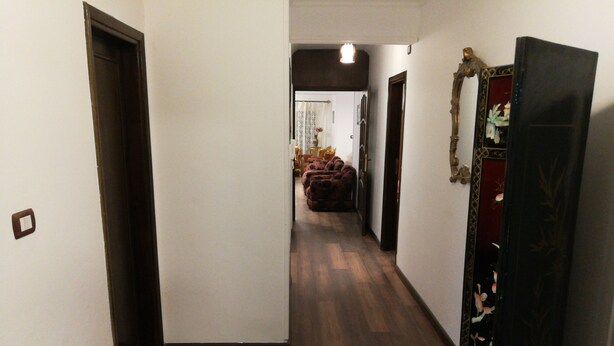 Een smalle gang met houten vloer, een deur links en een decoratief paneel rechts.