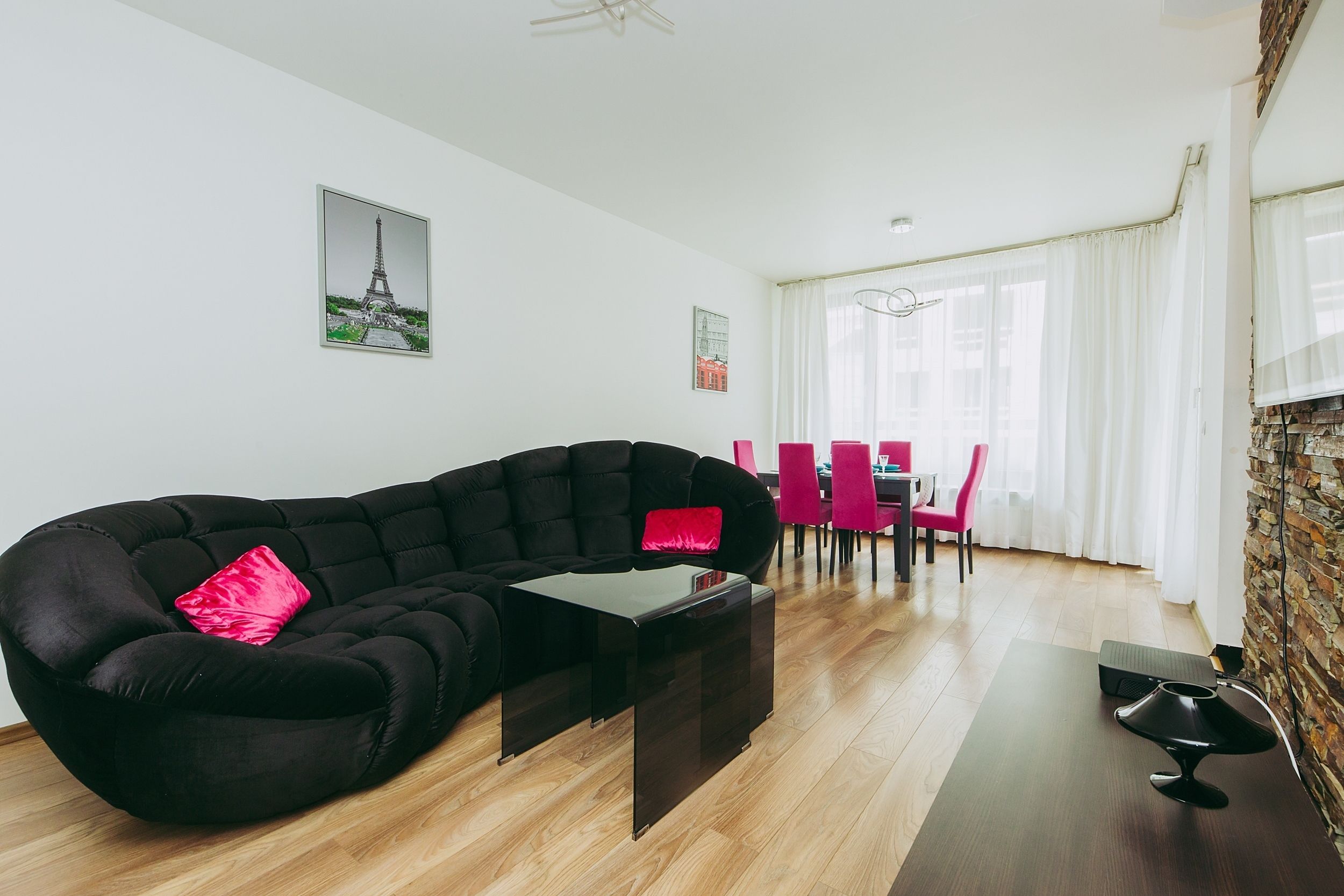 A modern living room with a black sectional sofa, a small black coffee table, and a dining area with pink chairs.