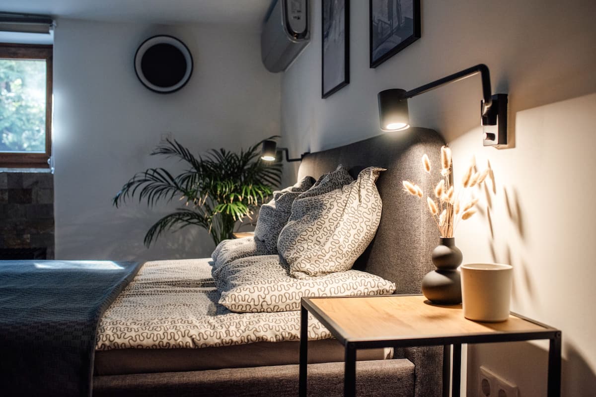 Ein Schlafzimmer mit einem Bett, einem Nachttisch, einer Lampe, einem Topfpflanze und einem Fenster.
