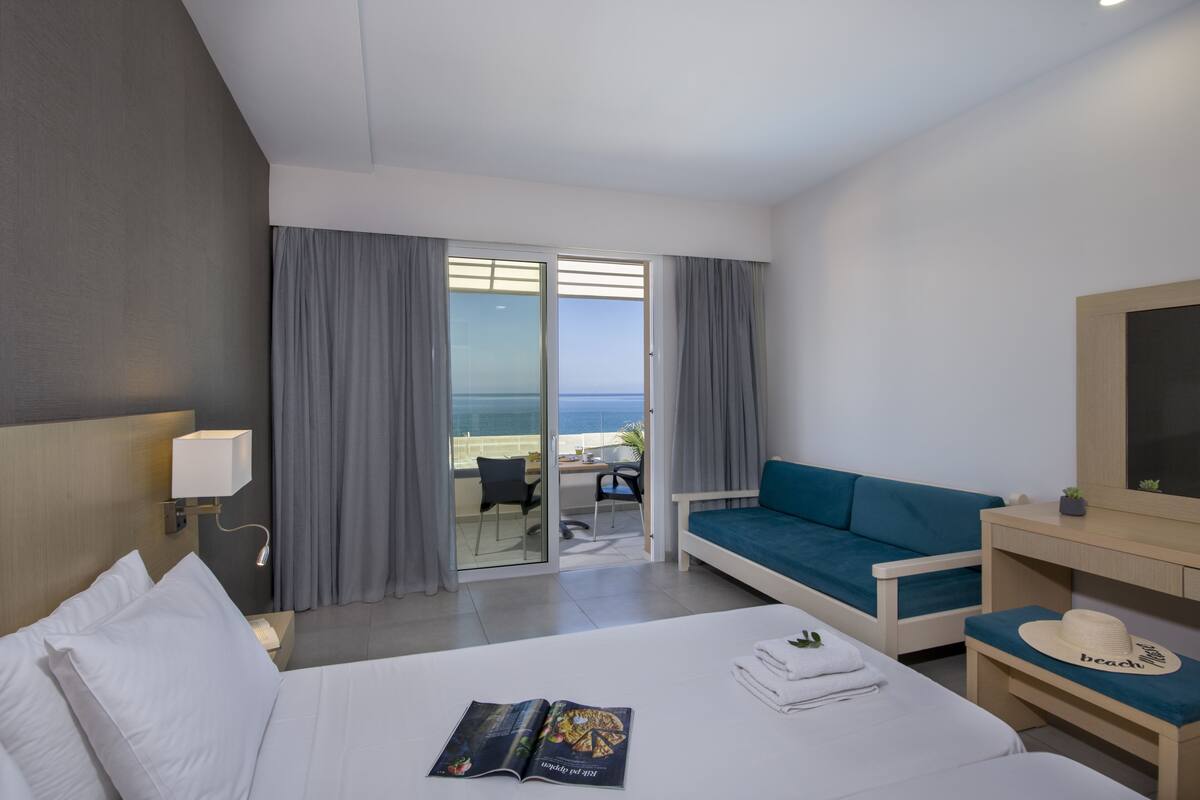 A hotel room with a bed, a sofa, a desk, and a view of the sea through a sliding door.