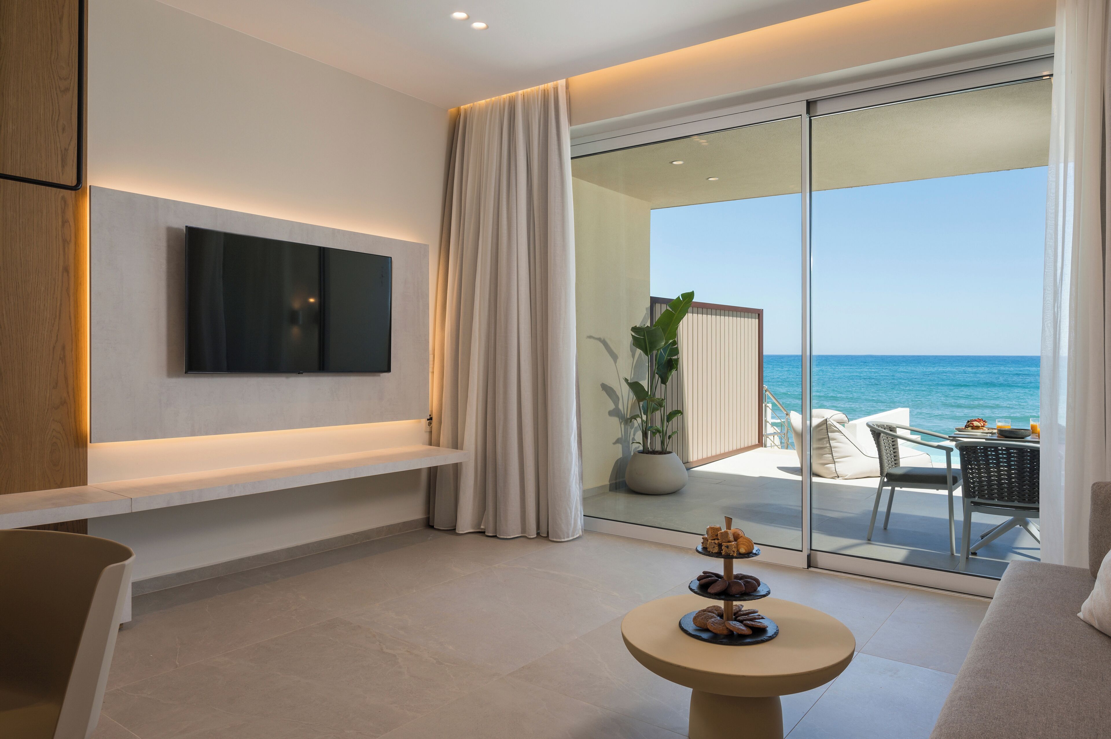 Une chambre d’hôtel moderne dotée d’un téléviseur à écran plat fixé au mur, d’un balcon offrant une vue sur la mer et d’une petite table avec des pâtisseries.