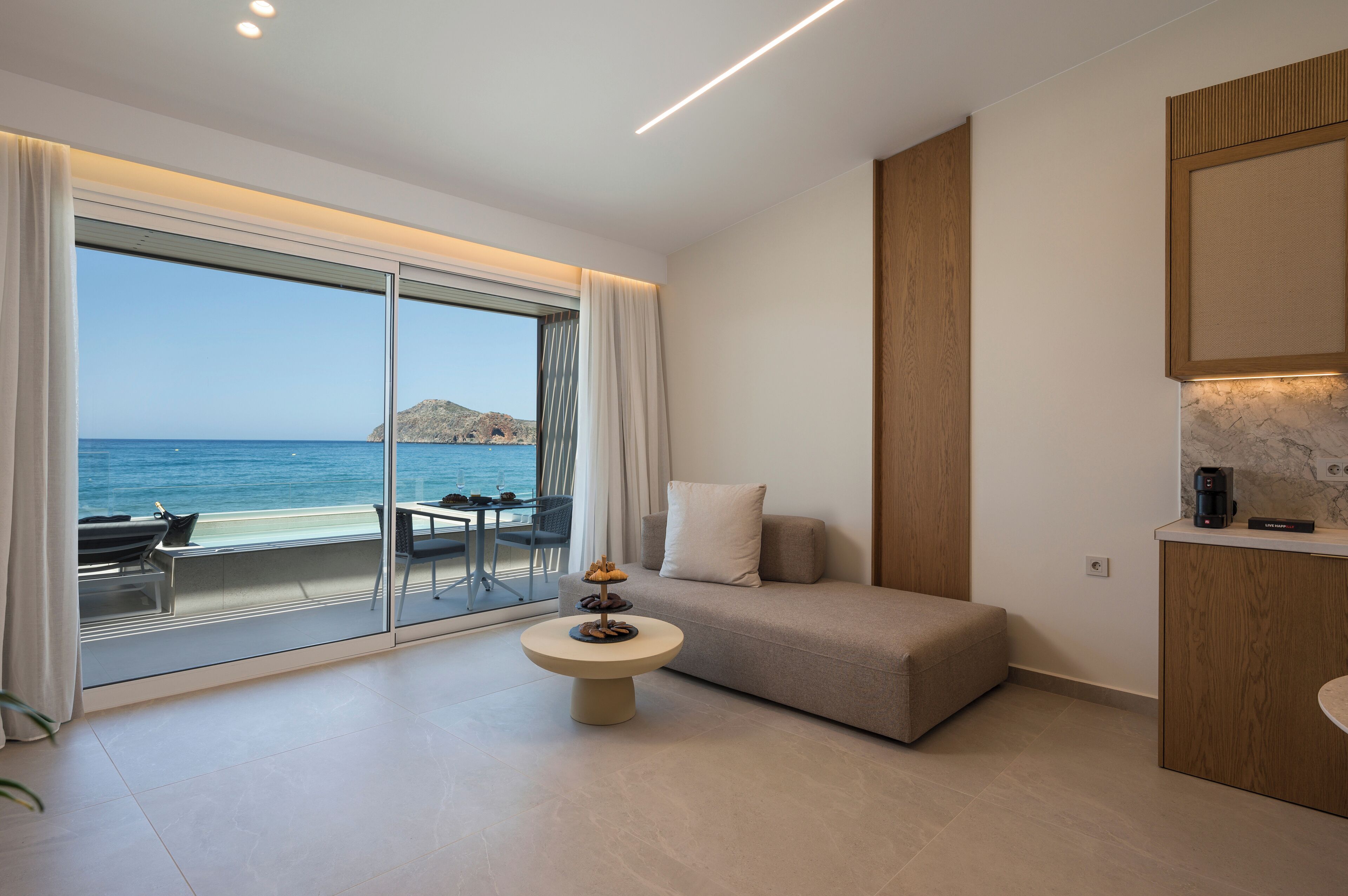 A modern hotel room with a large sliding glass door leading to a balcony with a view of the sea. There is a sofa, a small round table, and a coffee machine on a counter.