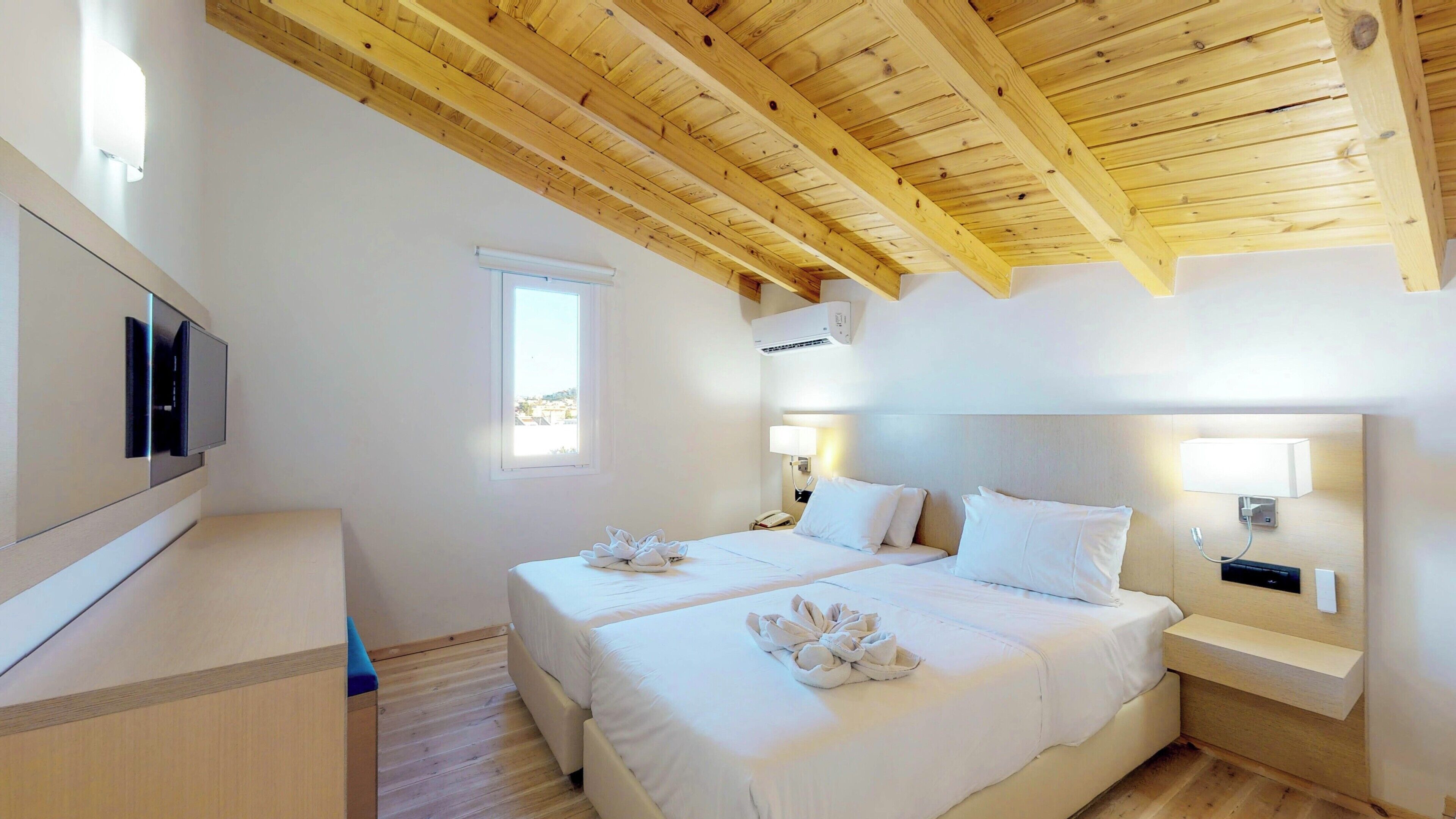 A hotel room with a wooden ceiling, two beds, a TV, a window, and a bedside lamp.