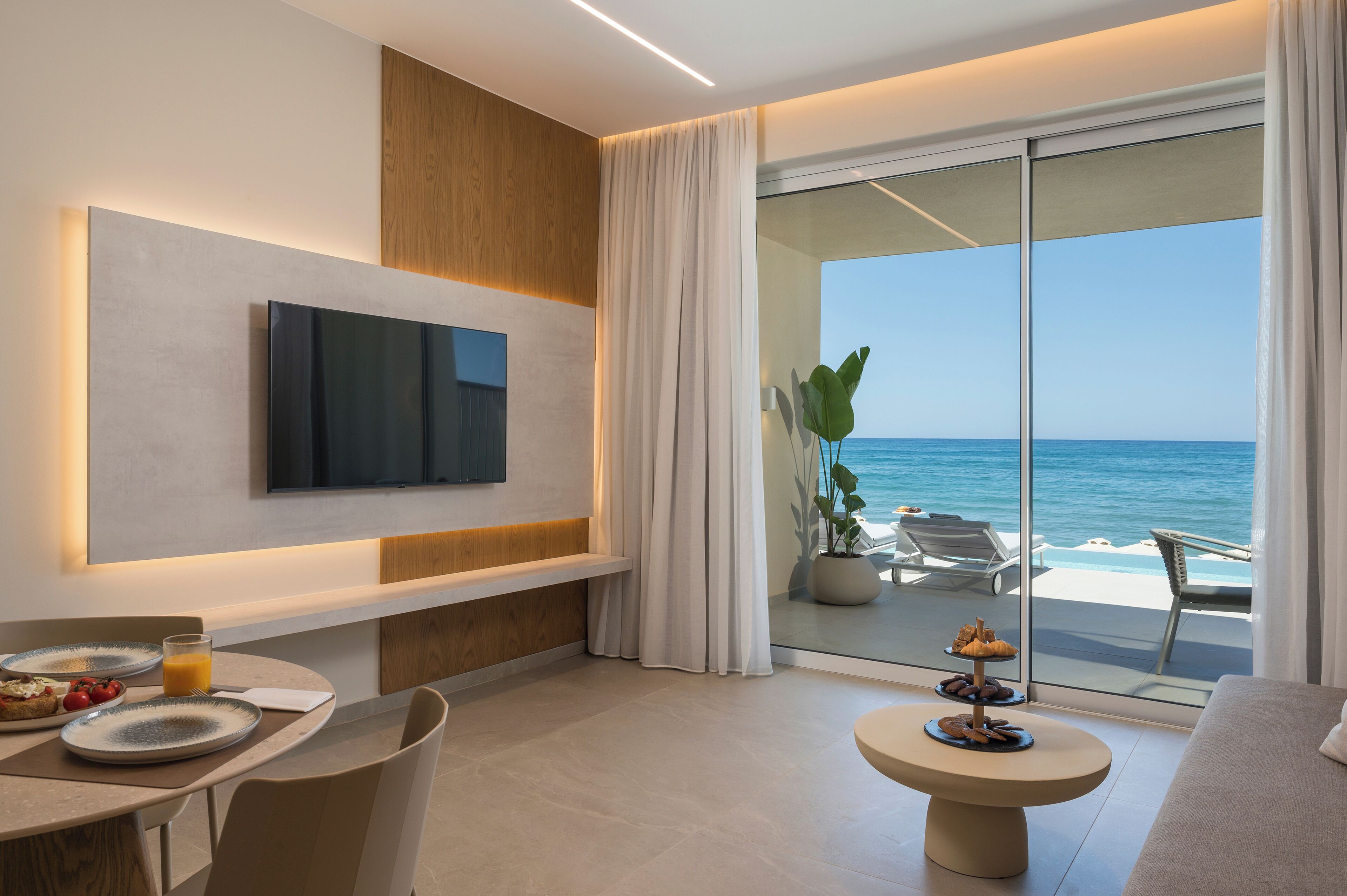 A modern hotel room with a large TV, a dining area with plates and a glass of orange juice, and a balcony with a view of the ocean.