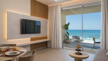 A modern hotel room with a large TV, a dining area with plates and a glass of orange juice, and a balcony with a view of the ocean.