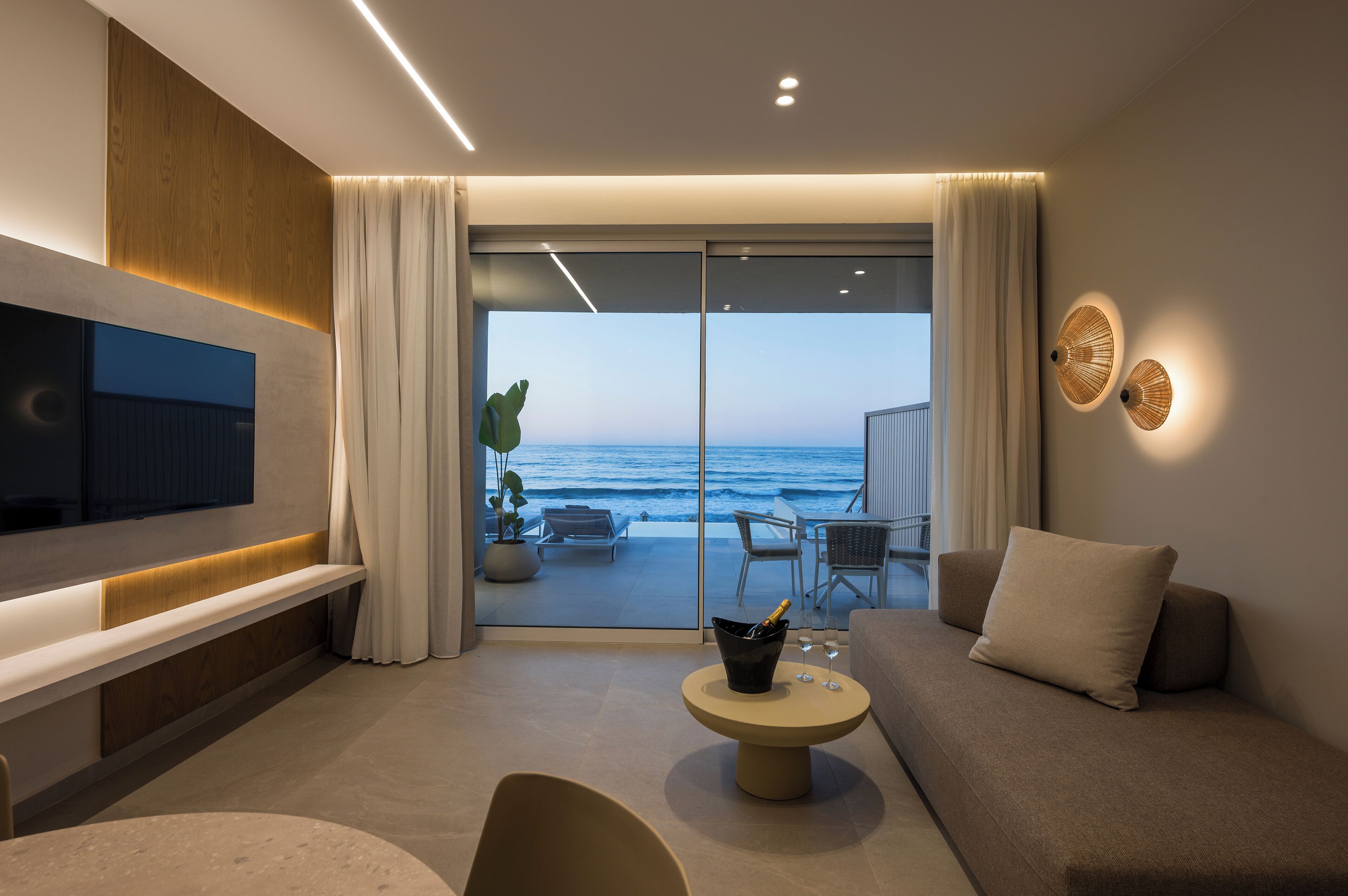 Uma sala de estar moderna com uma televisão grande, um sofá, uma mesa de centro com uma bebida e vista para o oceano através de grandes portas de vidro.