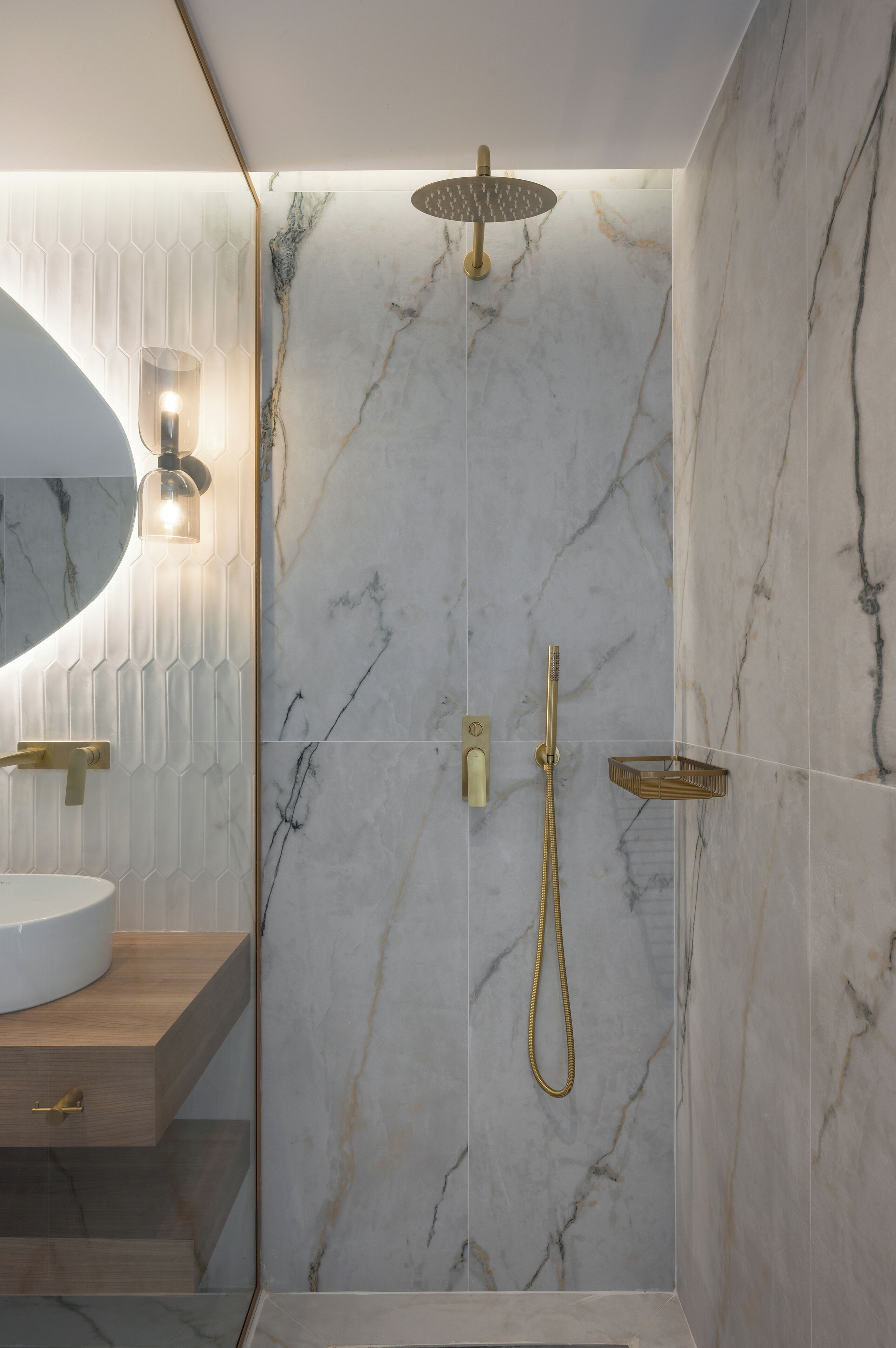 A modern bathroom with a marble shower wall, a wooden vanity, and a round mirror with a light fixture.