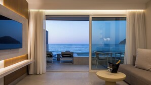 Habitación de hotel moderna con balcón y vistas al mar, televisión de pantalla plana en la pared, sofá, una mesita redonda con una botella y un vaso, y un par de sillas.