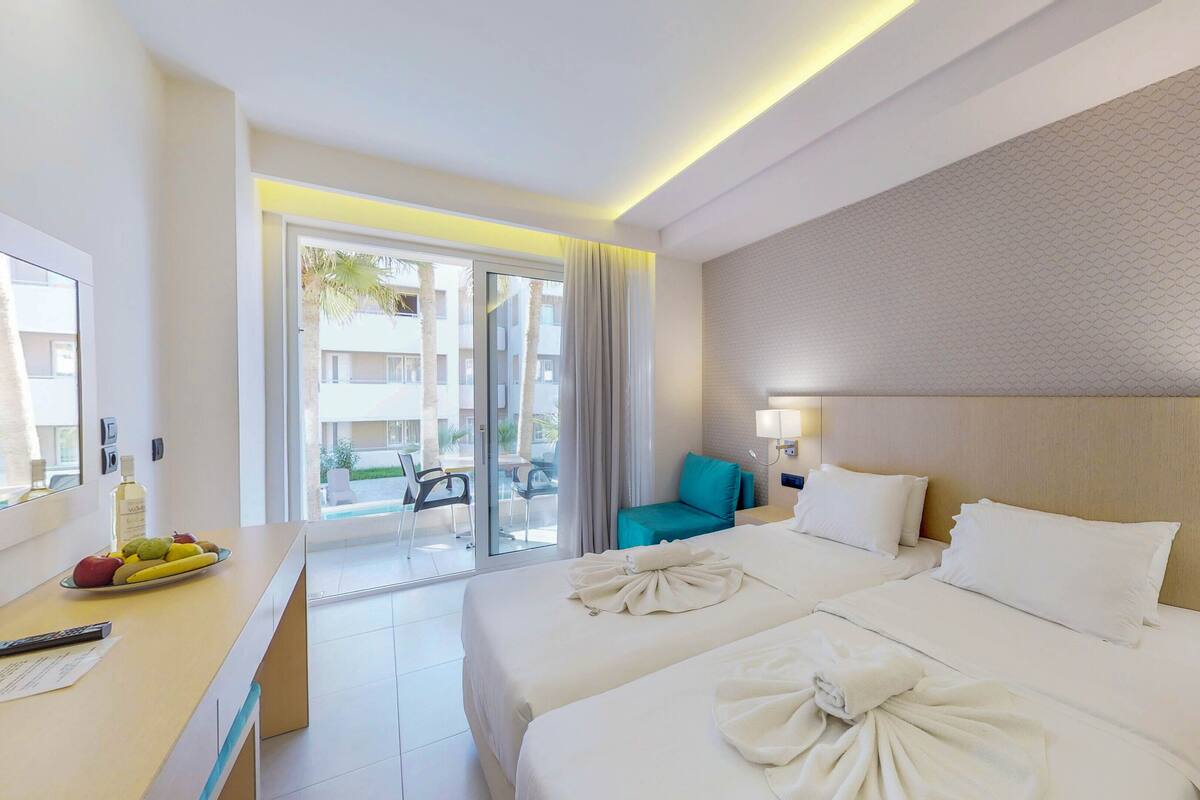 A hotel room with a large bed, a desk with a fruit bowl, a chair, and a view of the outside balcony.