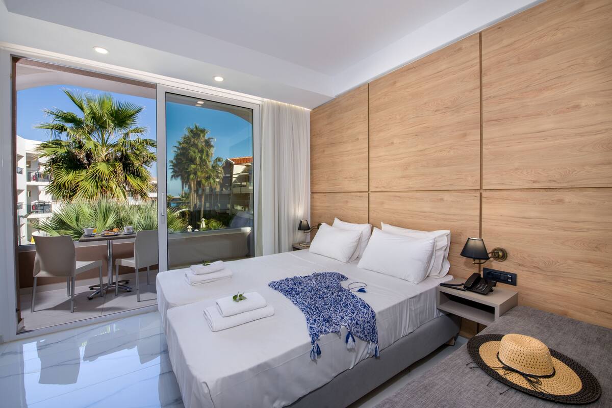 A modern hotel room with a large bed, a desk, and a view of palm trees through a sliding glass door.