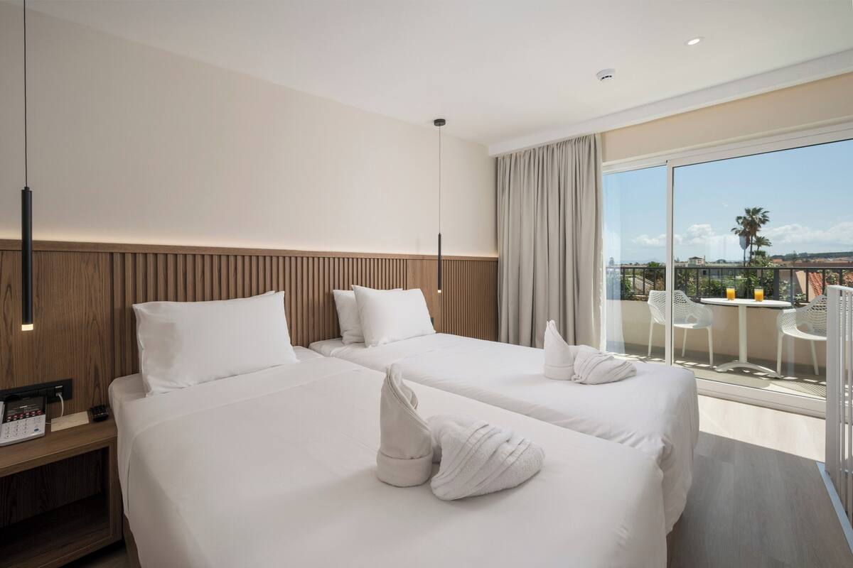 A hotel room with two beds, a wooden headboard, a nightstand with a phone, and a balcony with chairs and a table.