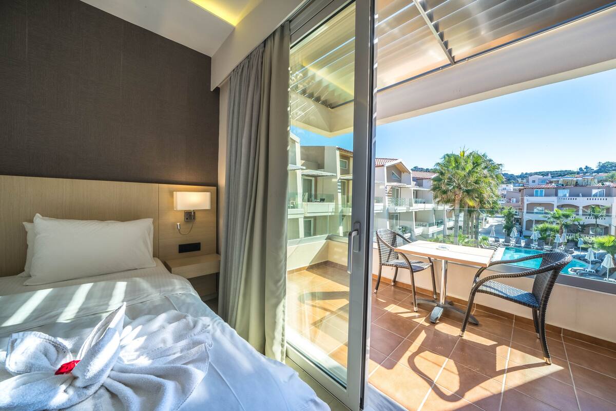 A hotel room with a bed, a glass door leading to a balcony with a pool and chairs.