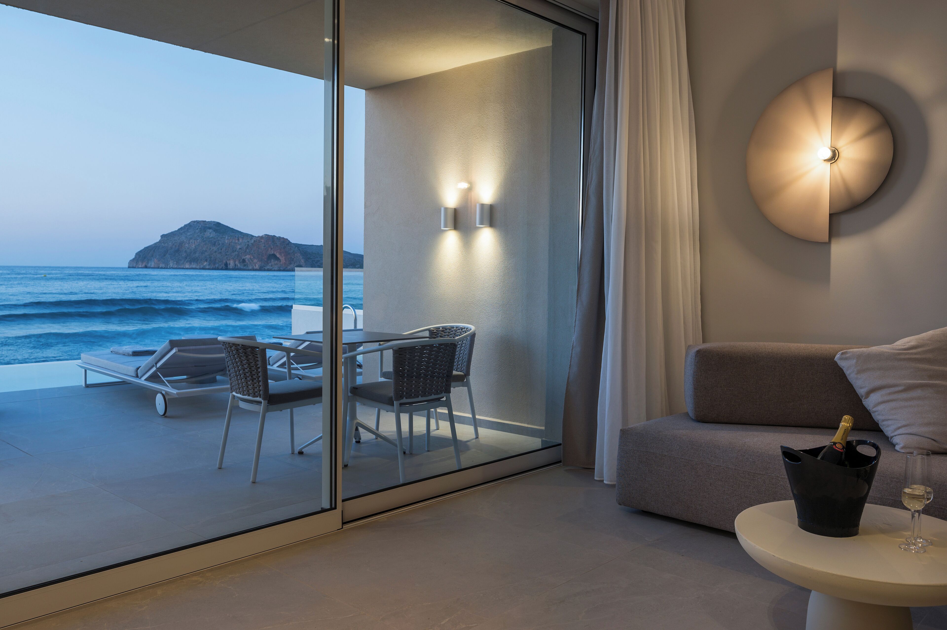 Un balcone con una porta a vetri, un tavolo con sedie, un divano, un lampadario a parete e una bottiglia di champagne.
