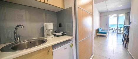 A compact kitchen with a sink, a coffee maker, and a mini-fridge. The room has a view of the sea through large windows.