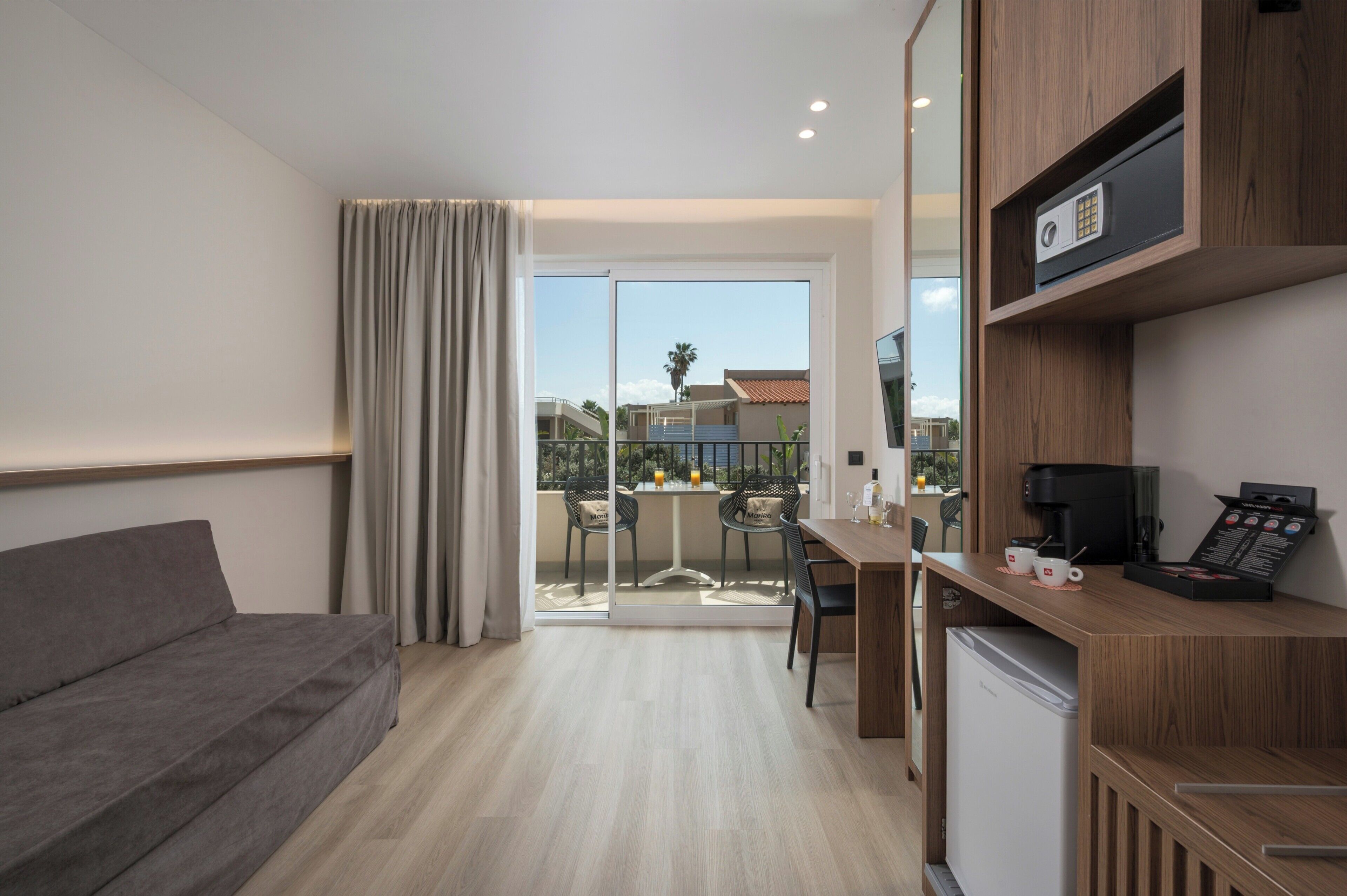 Habitación de hotel moderna con sofá, zona de comedor con mesa y sillas, cocina equipada con cafetera y puerta corrediza de vidrio que da a un balcón.