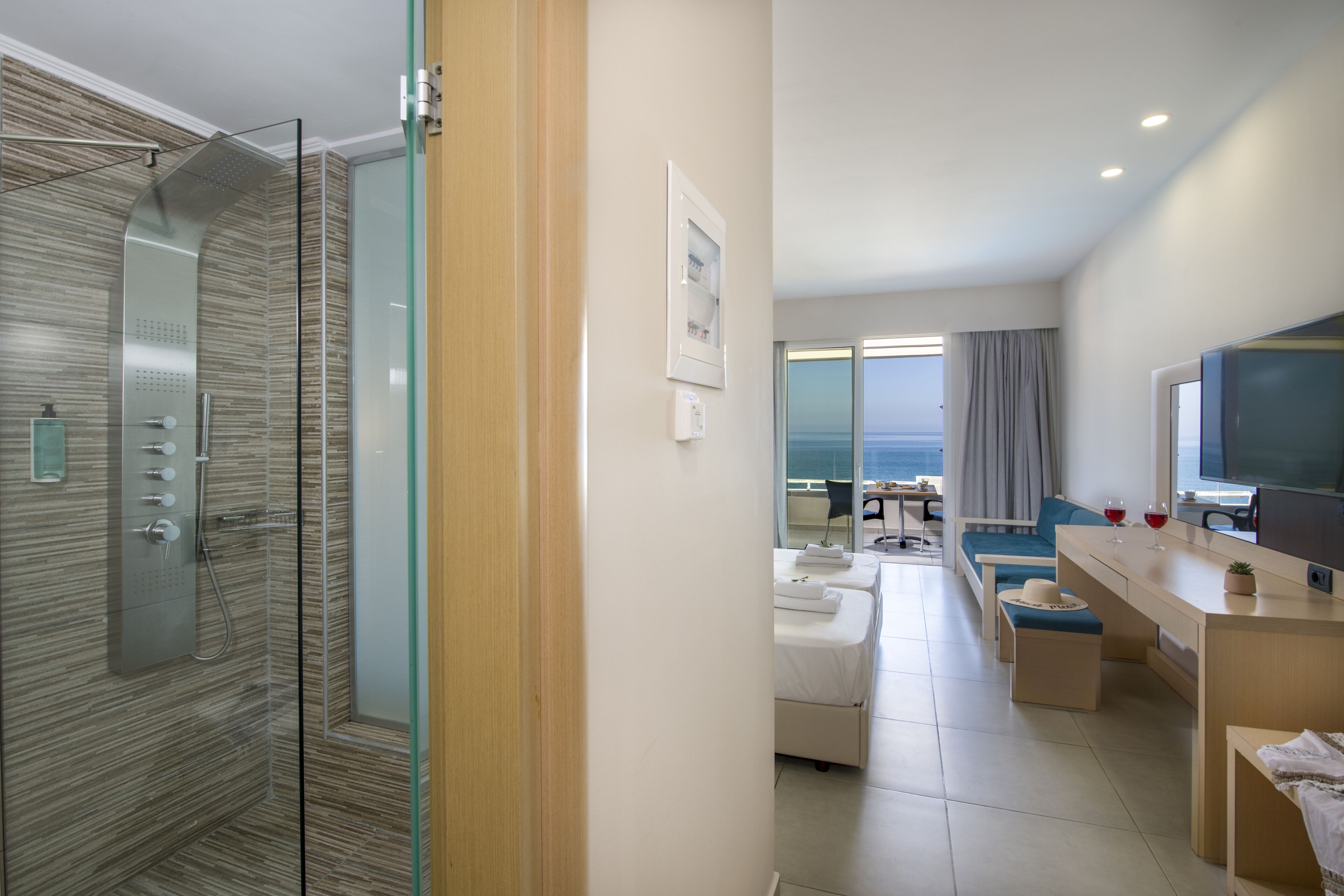 A modern hotel room with a glass-enclosed shower, a seating area with a sofa and chairs, a dining table, and a television.