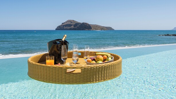 Una cesta flotante con frutas, bebidas y una botella junto a una piscina azul y despejada.