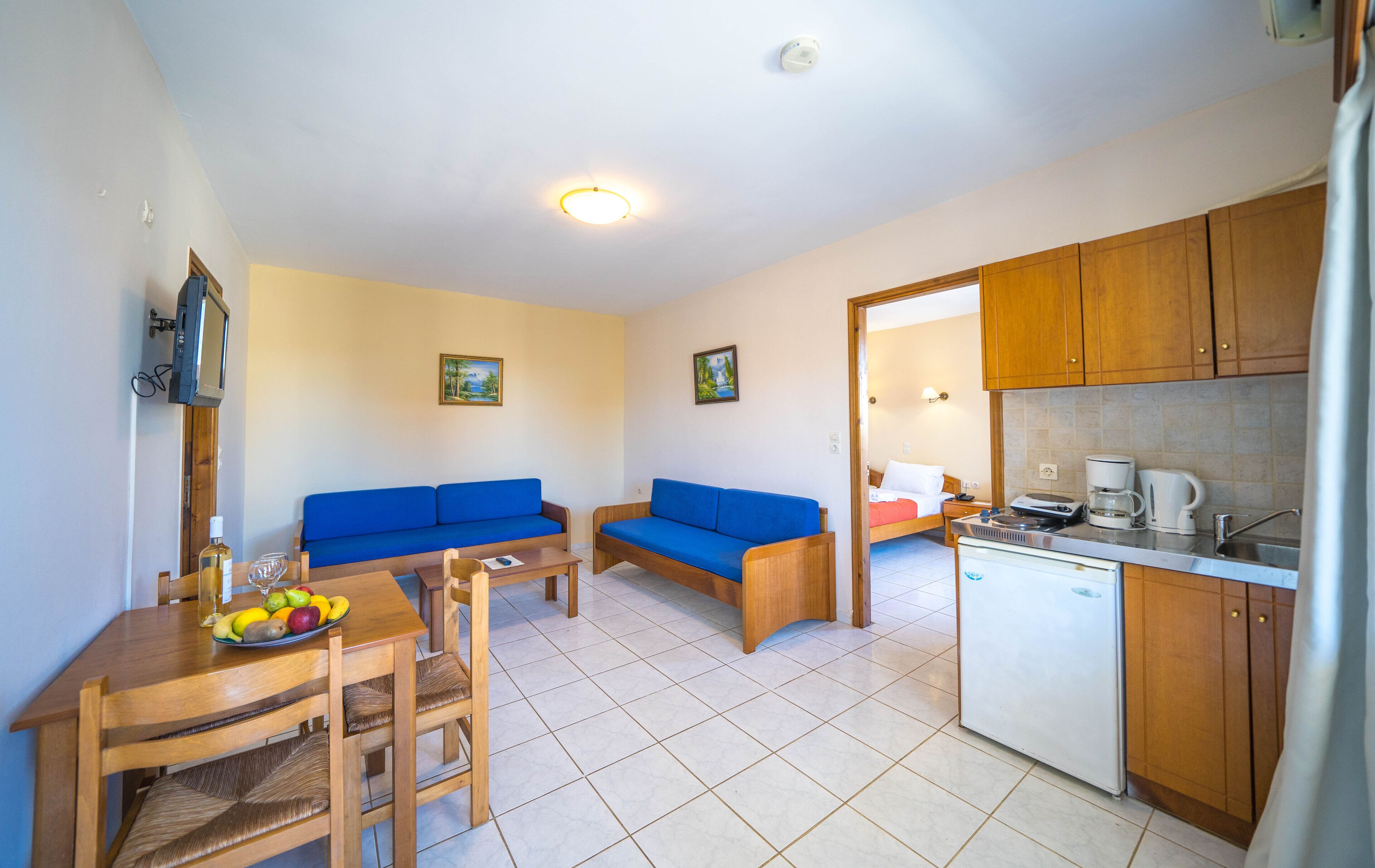 A compact living space with a sofa, a dining table with chairs, a kitchen area with a refrigerator, and a television mounted on the wall.