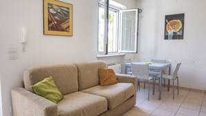 Ein Wohnzimmer mit einem beigen Sofa, zwei Kissen, einem Esstisch mit Stühlen, einem Fenster mit Jalousien und einem an der Wand montierten Telefon.