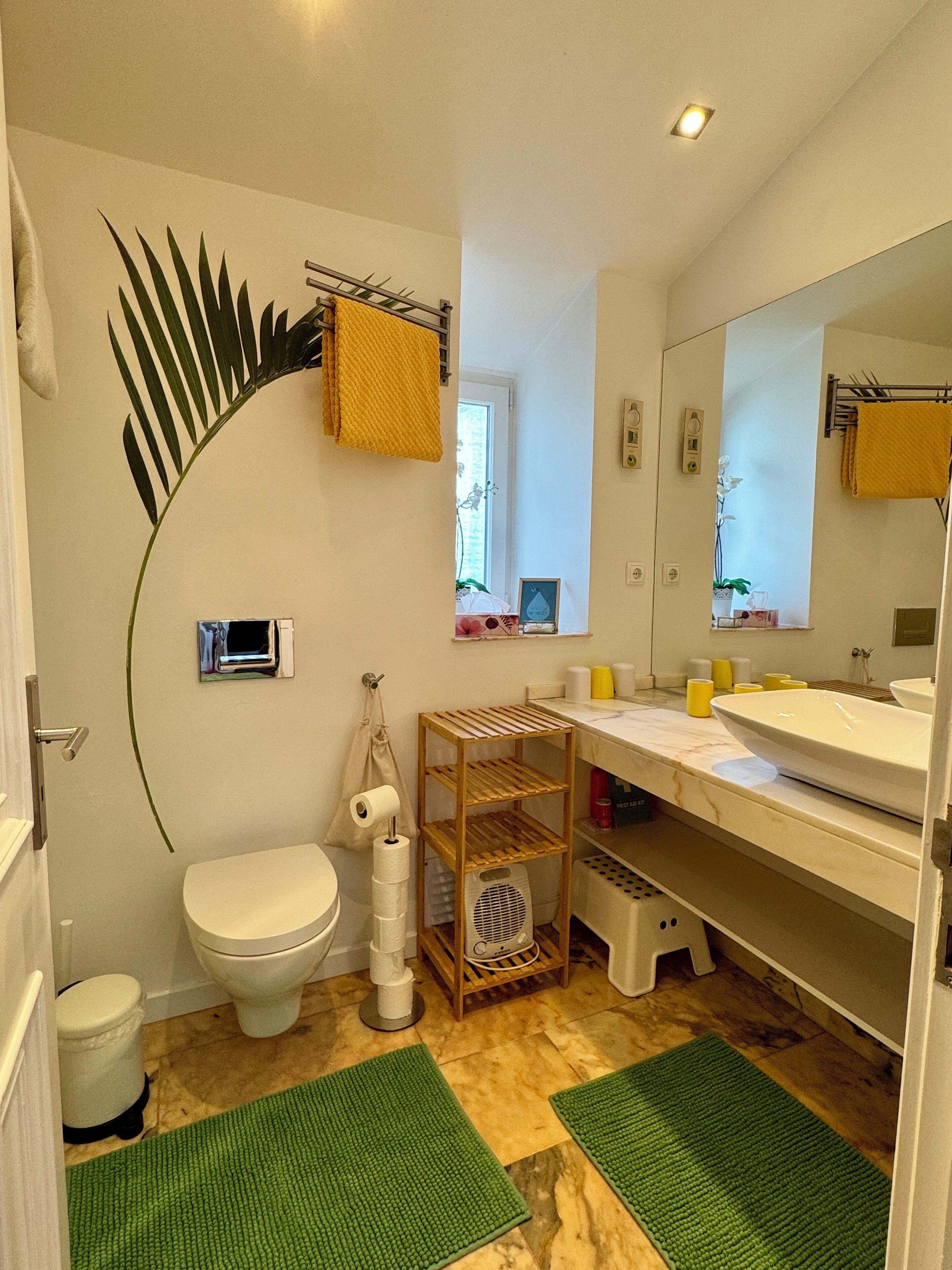 Et badeværelse med toilet, vask, spejl og et hyldesæt med toiletartikler.