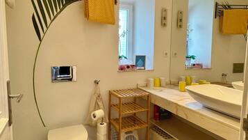 Et badeværelse med toilet, vask, spejl og et hyldesæt med toiletartikler.