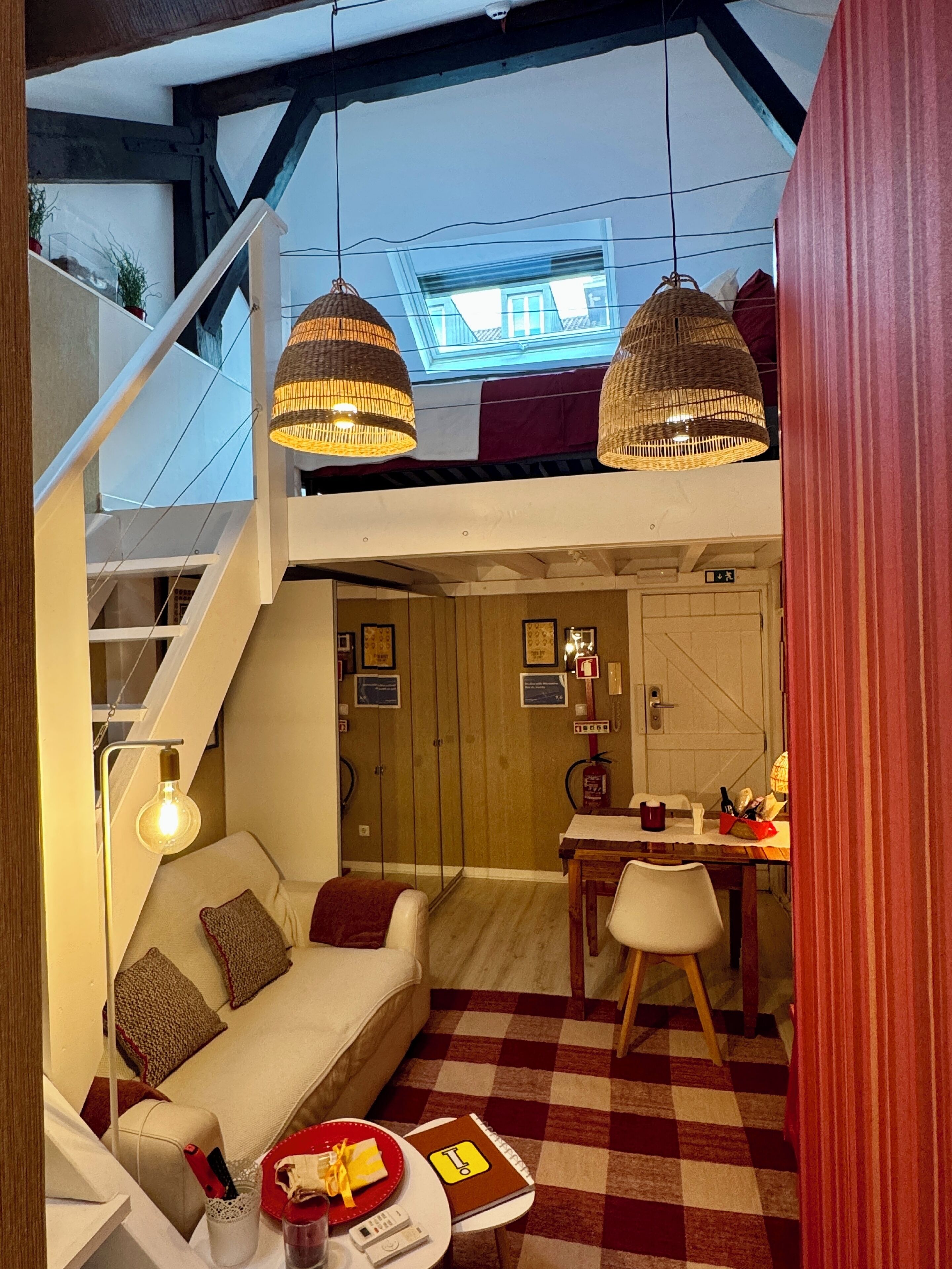Uma sala de estar aconchegante com um sofá, uma mesa de jantar e piso xadrez. Há luminárias de vime penduradas no teto e uma escada que leva a um andar superior.