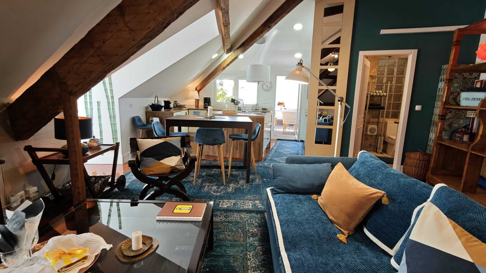 A cozy living room with a blue sofa, wooden beams, a dining area, and a kitchen in the background.
