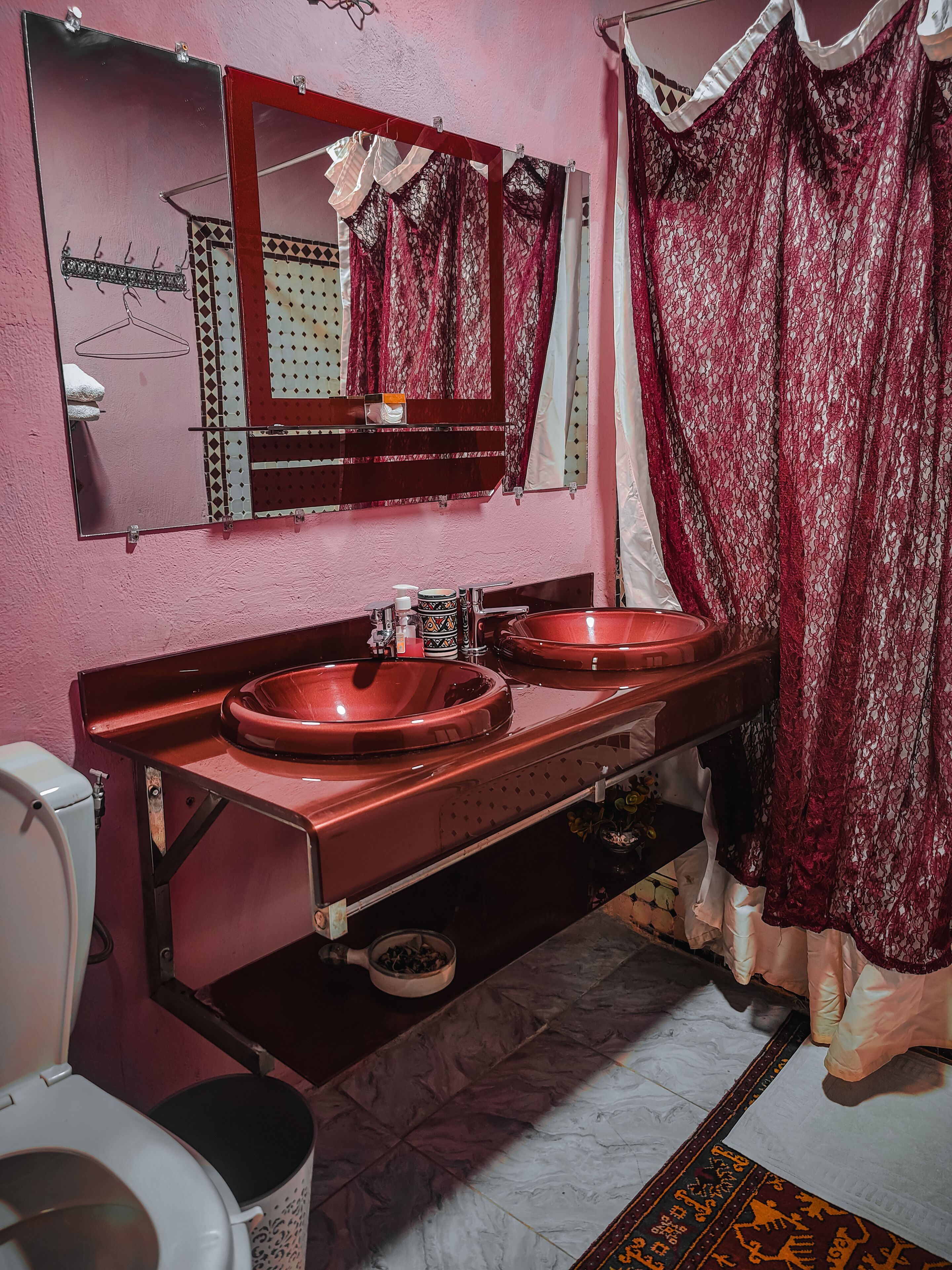 Une salle de bain avec un mur rose, un miroir, deux lavabos, des toilettes et un rideau de douche.