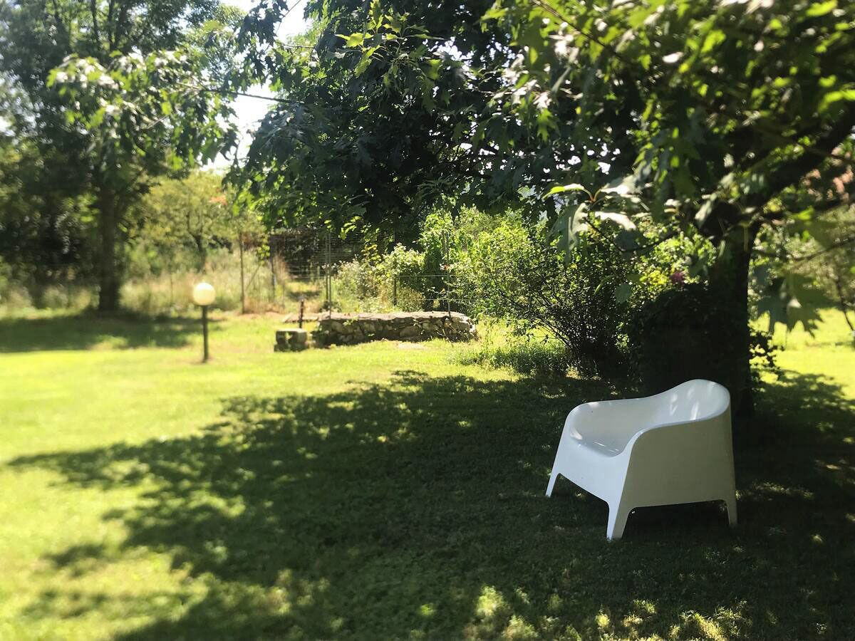 A white plastic chair in a grassy outdoor area with trees and a stone wall in the background.