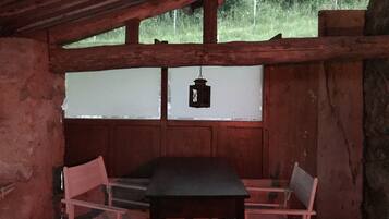 Una stanza rustica in legno, con un tavolo, due sedie, un lampadario appeso e una finestra che si affaccia sul verde.