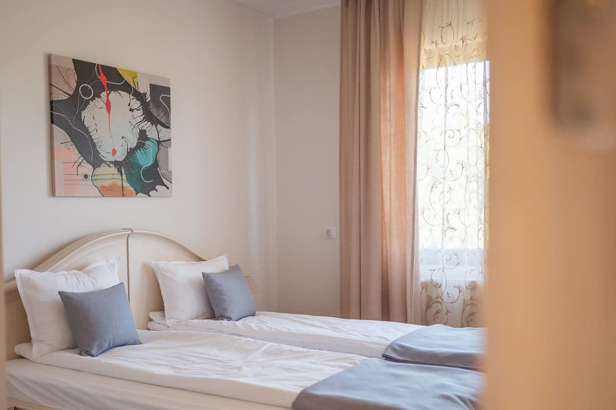 A bedroom with two beds, a painting on the wall, and a window with curtains.