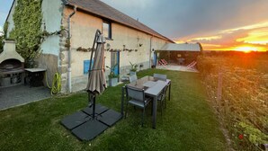 A rustic stone building with a sloped roof, a patio with a table and chairs, and a view of the sunset. - Charming independent cottage with enclosed garden, pets allowed (La Petite-Marche)