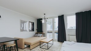 Ett modernt hotellrum med en säng, en soffa, ett matbord med stolar, en liten bänk och utsikt över staden genom stora fönster.