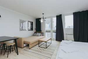 Una habitación de hotel moderna con cama, sofá, mesa de comedor con taburetes, un banco pequeño y vistas a la ciudad a través de amplias ventanas.