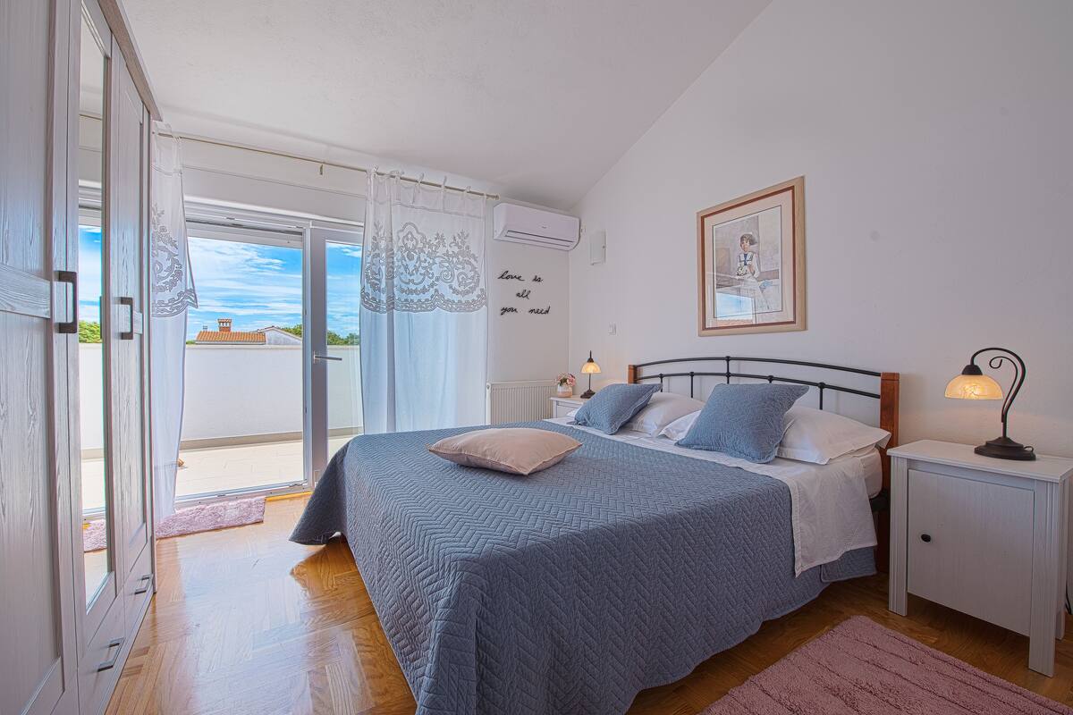 A bedroom with a bed, a nightstand, a lamp, a framed picture, and a window with a view of buildings.