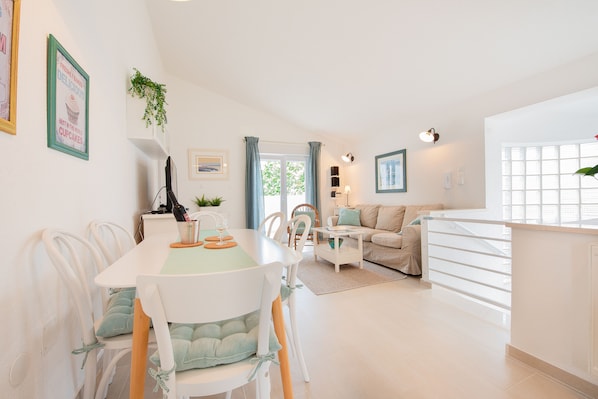 A modern, well-lit living area with a dining table, chairs, a sofa, and a kitchen area.