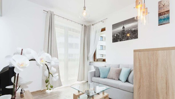 A modern living room with a glass coffee table, a sofa with cushions, a wooden sideboard, and a large window with curtains. - Modern Apartment | Parking | Close to Tauron Arena (Kraków)