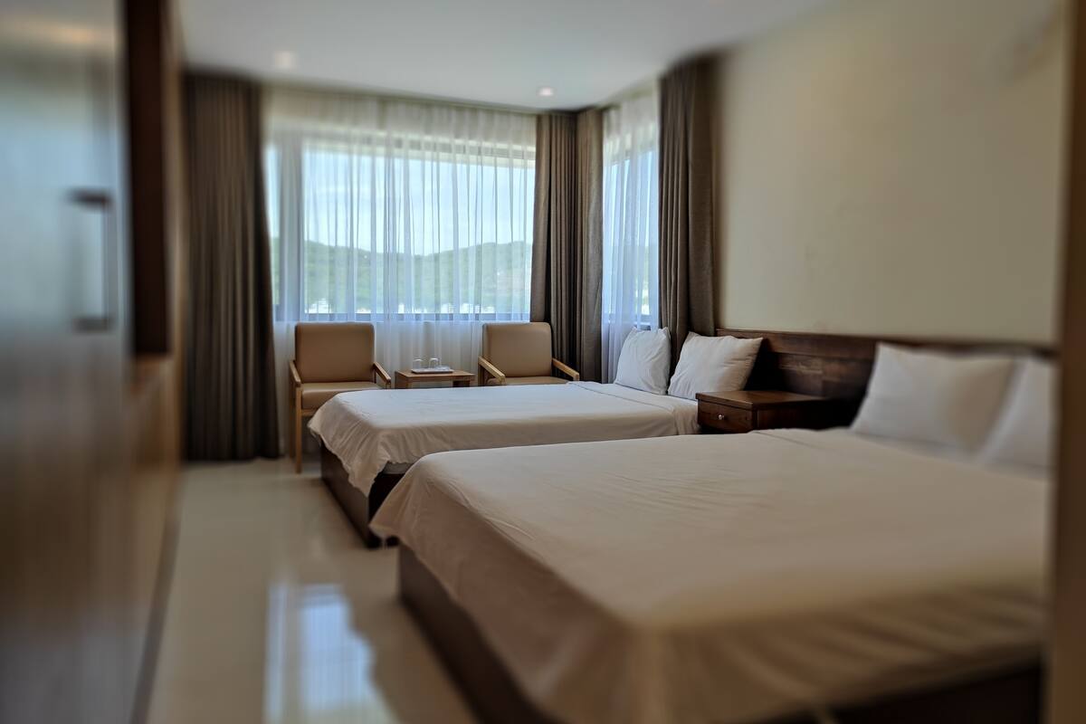 A hotel room with two beds, a sitting area with chairs, a small table, and a view of the outdoors through large windows.