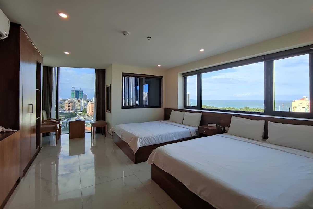 A modern hotel room with two beds, a large window with a city view, a desk, and a wardrobe.