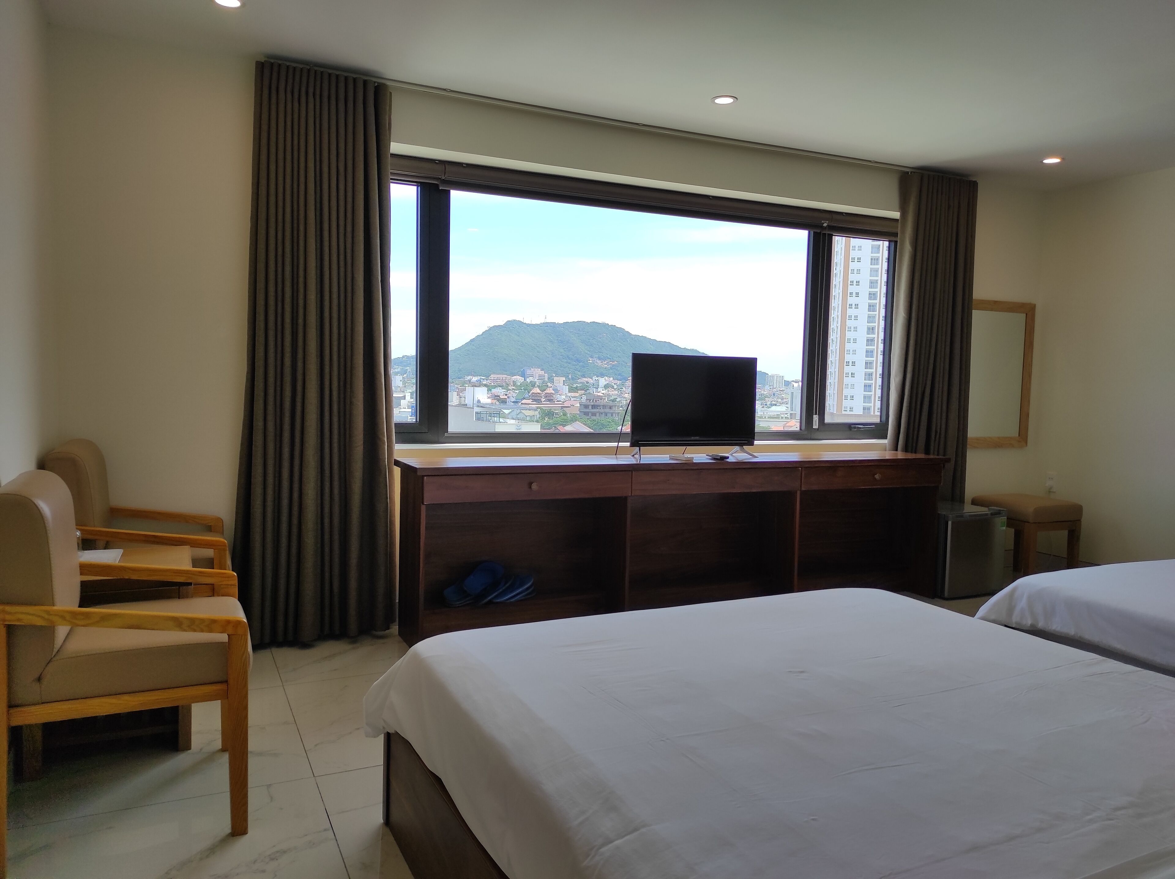 A hotel room with a bed, a desk with a TV, a chair, and a view of a cityscape and mountains.