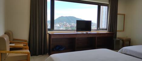 A hotel room with a bed, a desk with a TV, a chair, and a view of a cityscape and mountains.