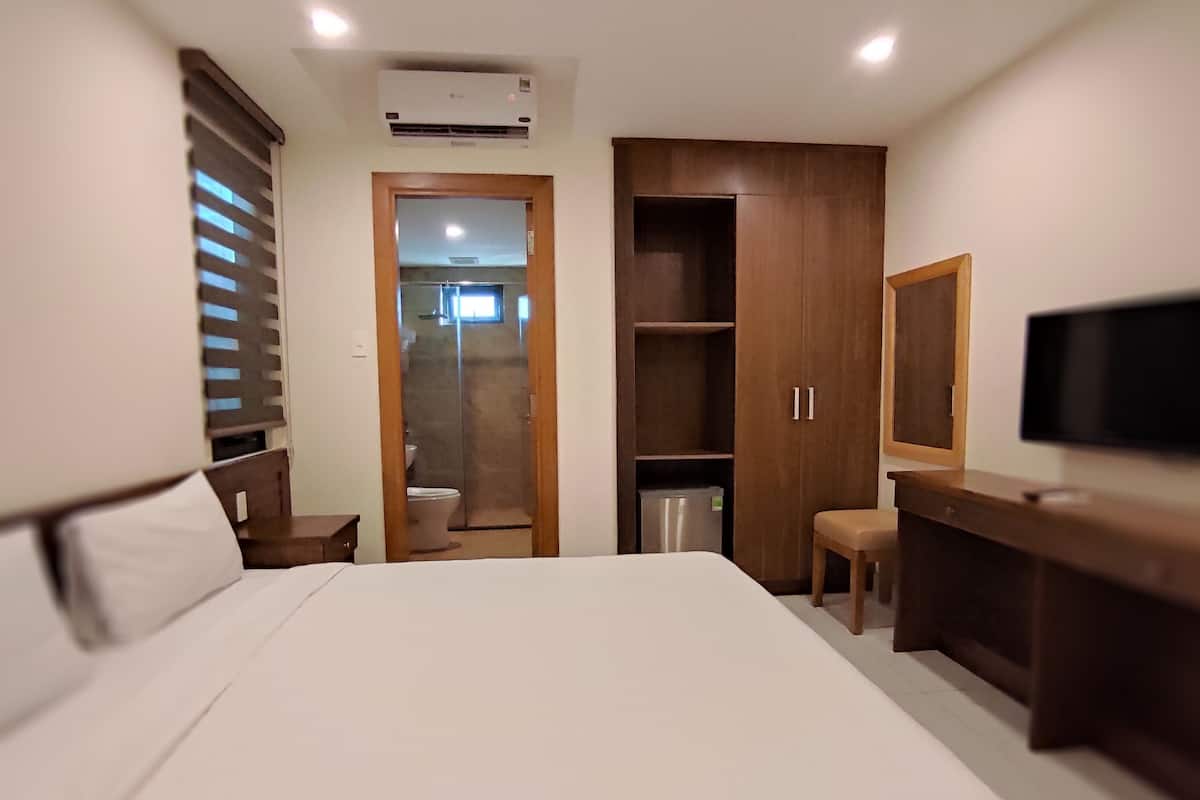 A hotel room with a bed, a wooden wardrobe, a desk with a chair, a television, and a bathroom visible through a door.