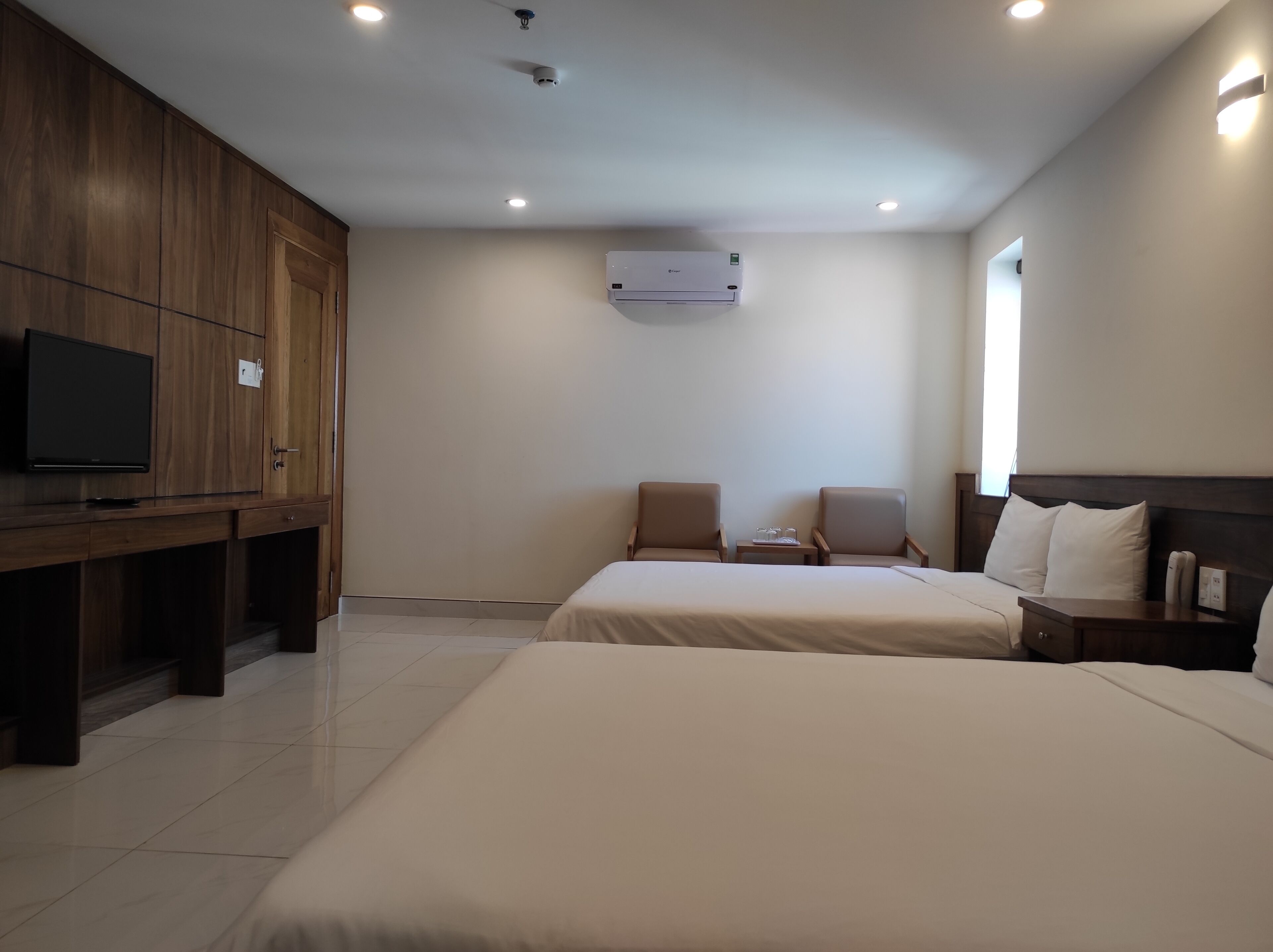 A hotel room with a bed, two chairs, a TV, and an air conditioning unit.