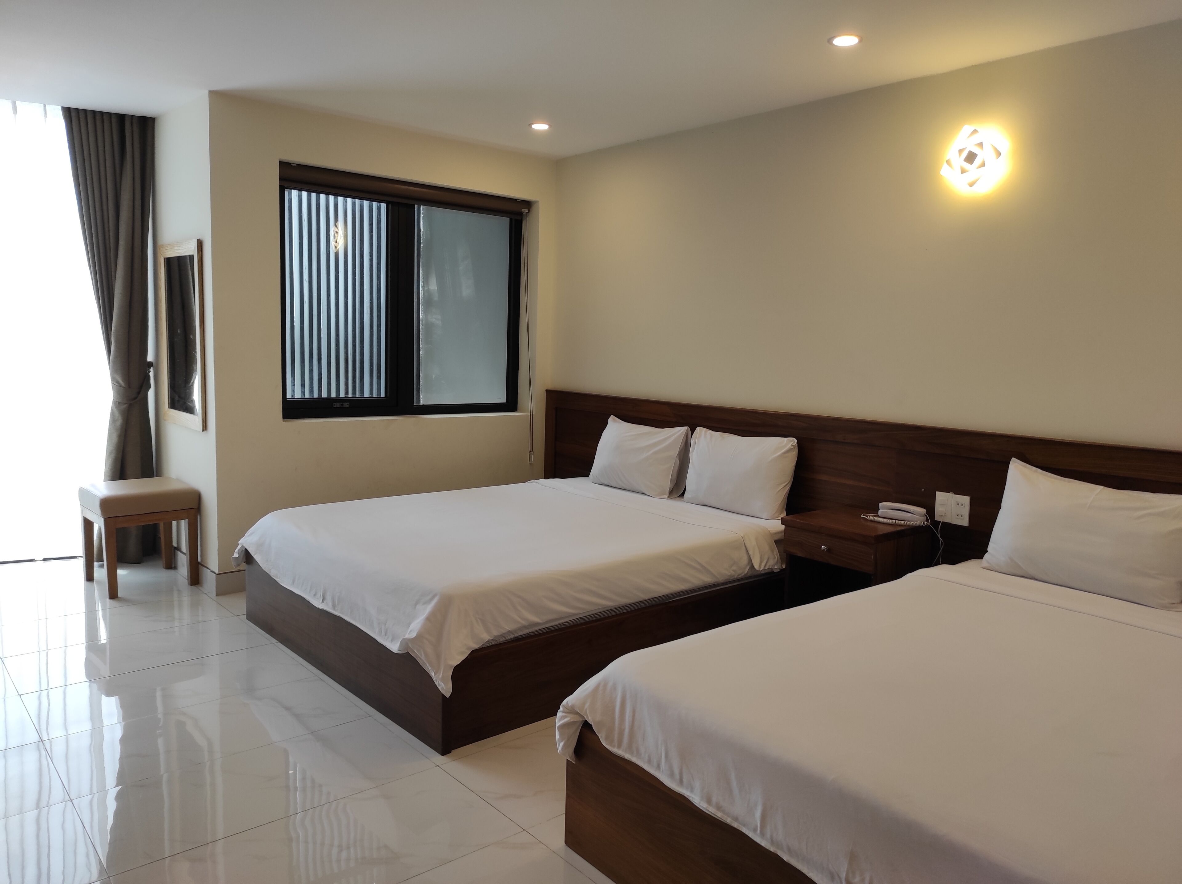 A hotel room with two beds, a small table, a window with blinds, and a wall-mounted light.