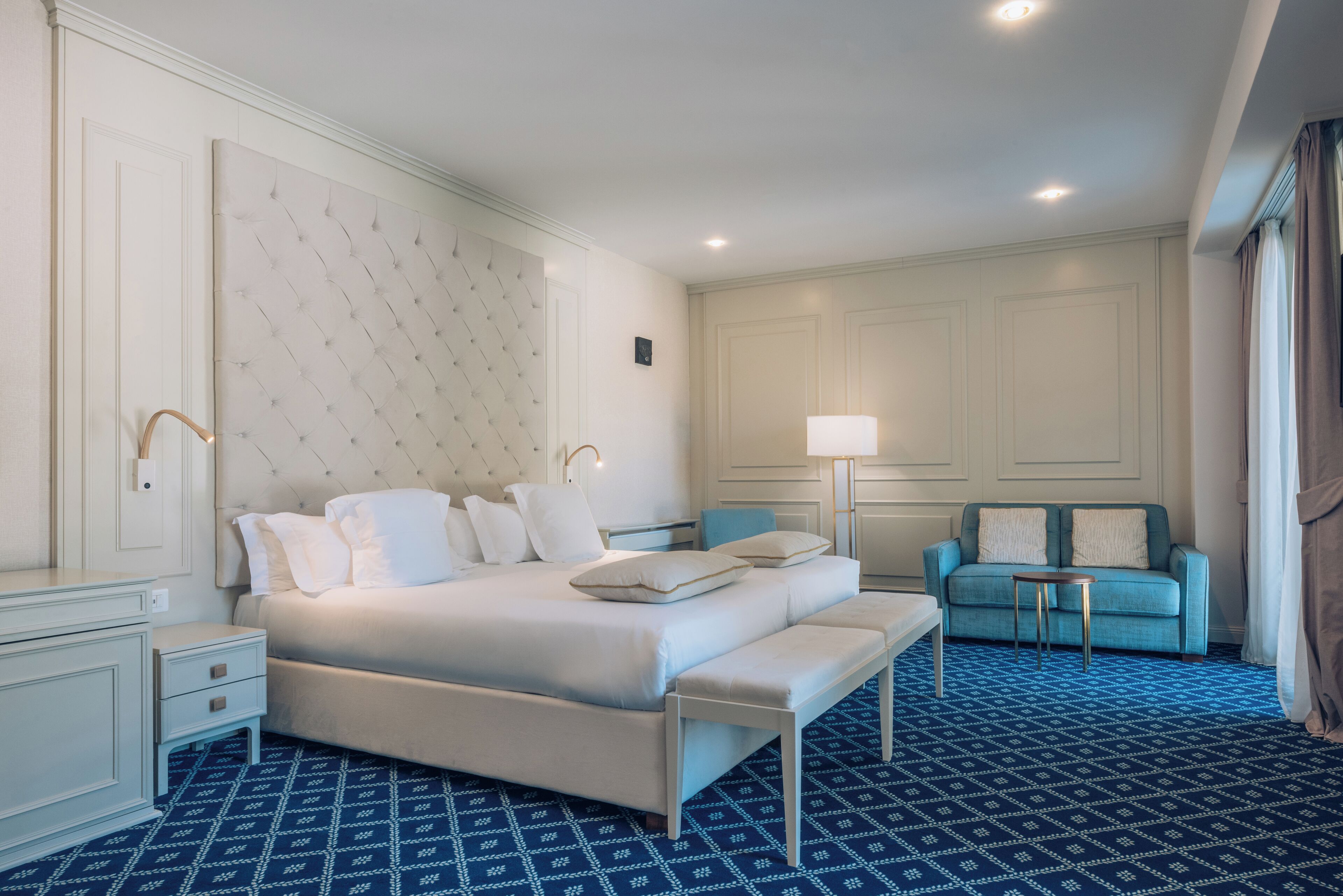A hotel room with a large bed, a bedside table, a bench, a blue patterned carpet, a blue sofa, and a lamp.
