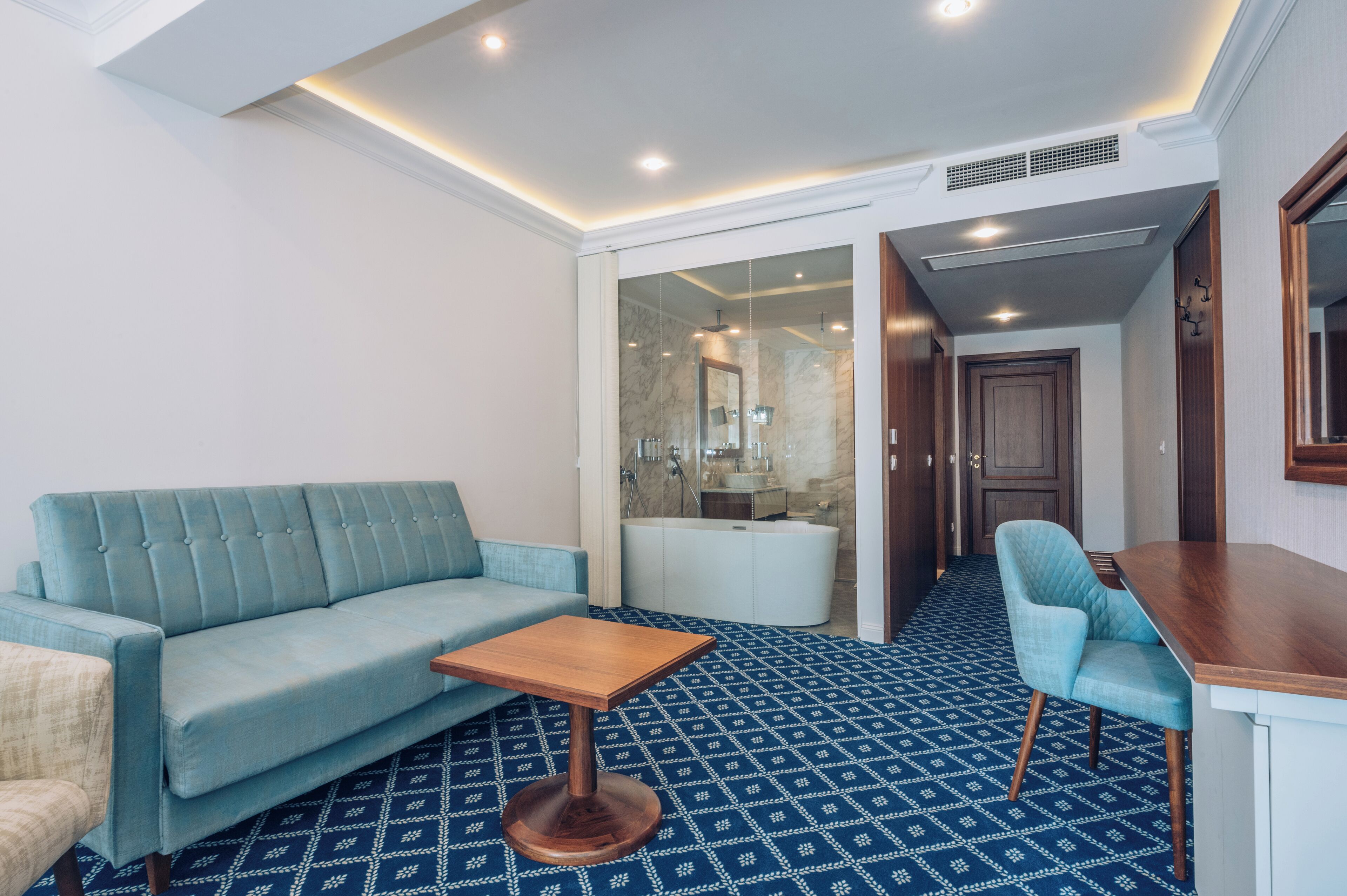Une chambre d’hôtel moderne avec un tapis à motifs bleus, un canapé bleu clair, une table basse en bois et une table d’appoint en bois.